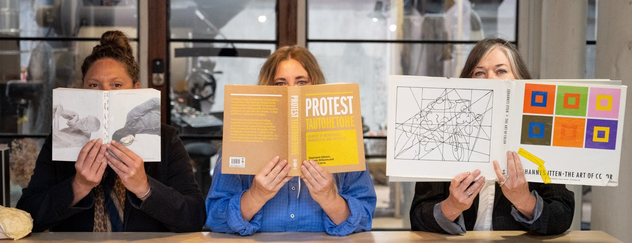 Three people sitting at a table holding books in front of their faces, with a modern interior background.